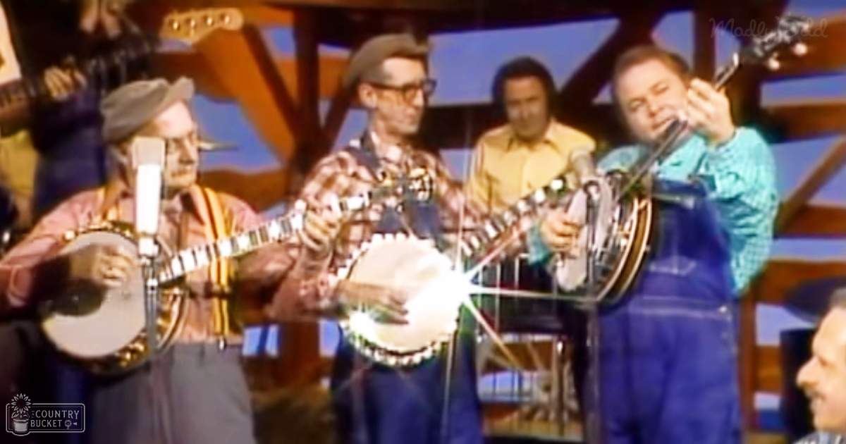 A group of adult males performing on a stage with microphones positioned in front of each person. The setting appears to be indoors with stage lighting highlighting the performers, and wooden structures are visible in the background.