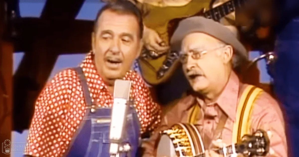 Musical performance with two adult male performers, one wearing a red polka-dot shirt and blue overalls, the other in a pinkish-red button-down shirt with yellow suspenders, on an indoor stage under dim lighting focused on them.