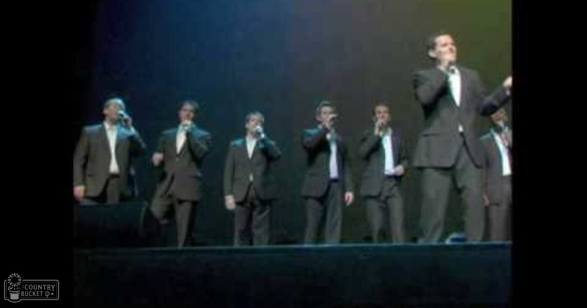 An adult male a cappella group performing on stage at an indoor venue, dressed in formal attire with microphones, under dim stage lighting.