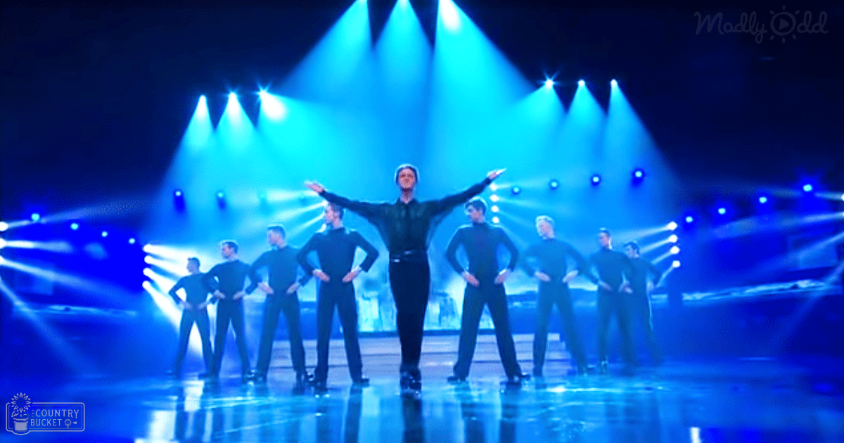An adult male dancer leads a line of young adult males in dark green shirts and black pants on a stage illuminated by blue spotlights.