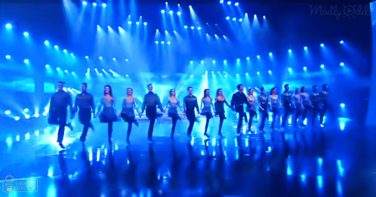 A group performance scene at an indoor stage event with young adults, both male and female dancers wearing traditional Irish dance attire under bright blue lighting on a reflective floor.