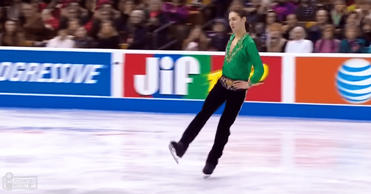 A young adult female figure skater in a green long-sleeved top and black pants, performing on an indoor ice rink with spectators watching.