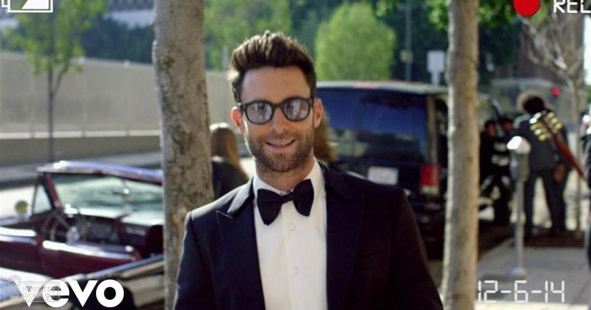 An adult male in a formal black tuxedo with thick-framed glasses stands outdoors near trees and parked cars as a band plays music, suggesting an outdoor event on a bright spring or summer day.