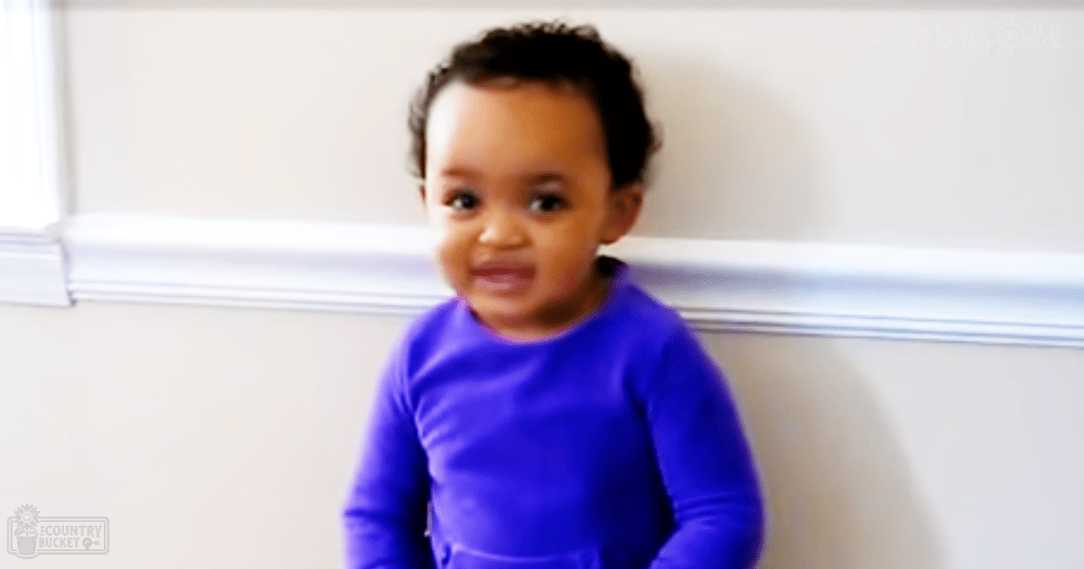 A toddler wearing a snug, long-sleeved solid purple garment indoors with natural light streaming in from a window.