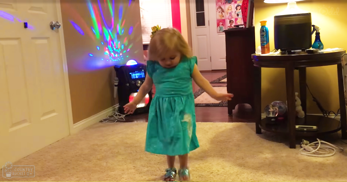 A toddler in a teal dress with shiny silver sandals stands indoors, surrounded by household items under artificial evening light.