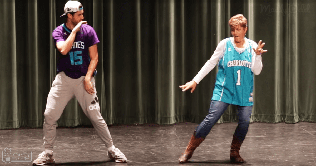 Adult male dancer in a purple Adidas basketball jersey with number 15, performing on an indoor stage with teal Charlotte Hornets jersey-wearing female companion.