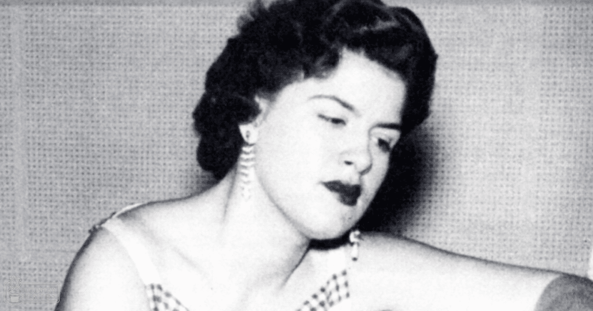 An adult female wearing pearl-like earrings and a checkered necklace indoors against a textured wall with grid pattern.