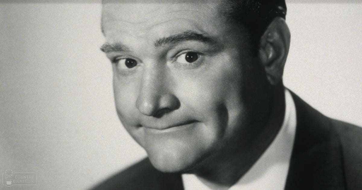 A middle-aged man wearing a dark suit jacket over a white collared shirt stands against a plain light-colored wall in what appears to be a studio setting with controlled lighting, typical of mid-20th century photography.