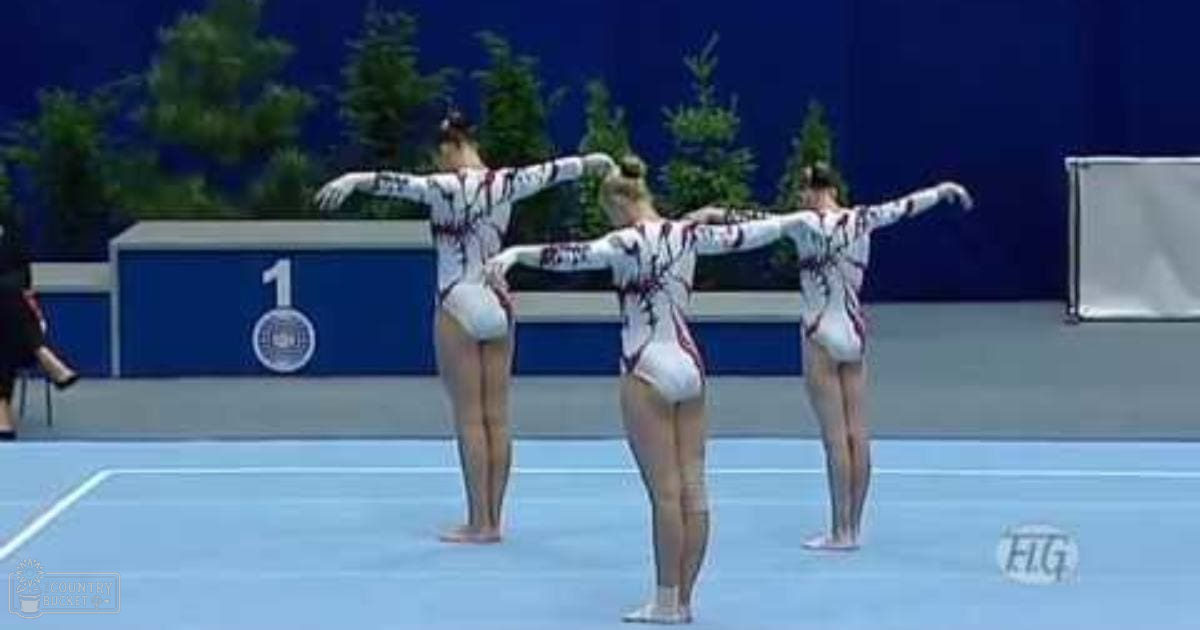 Young female gymnasts performing in an indoor arena with blue floors, walls, and greenery visible through a wall opening. They are wearing white leotards with red and black patterns.