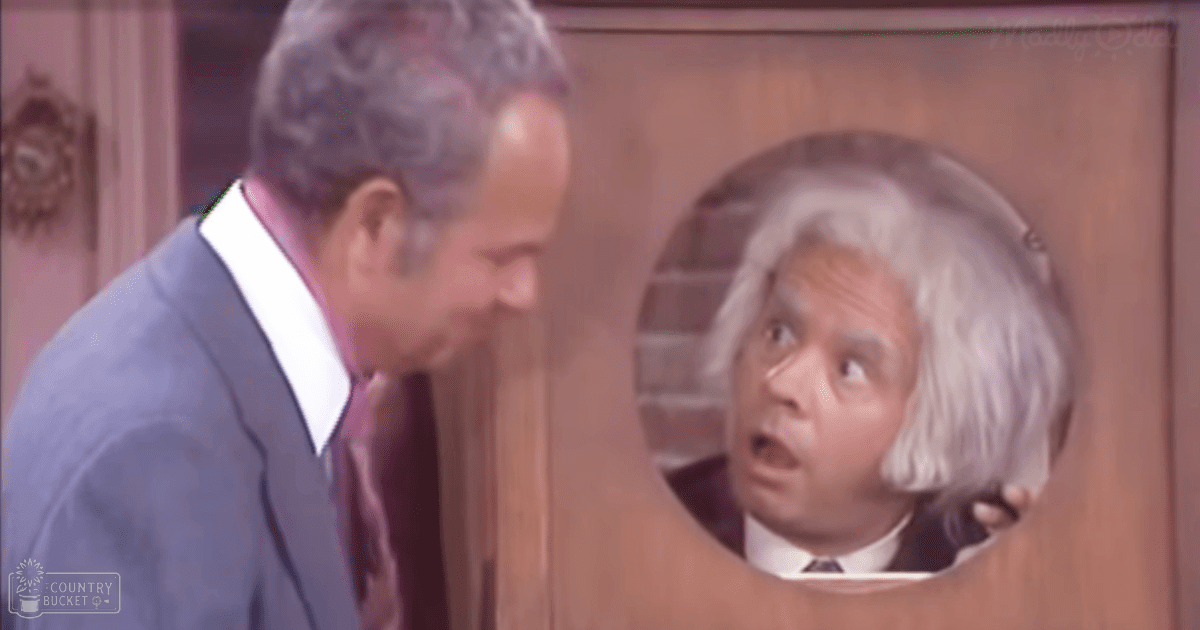 An older adult male in a dark blue suit jacket with a white shirt, maroon tie, and an older adult man wearing a white wig and dark suit, set against a wooden door indoors.