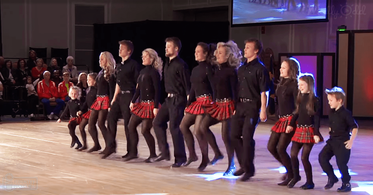 A dance performance in an indoor setting with children wearing black tops and red plaid skirts, adults dressed casually, and a large audience seated on chairs.