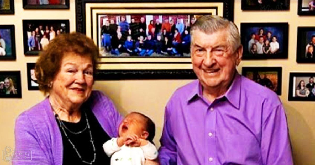 An older adult woman holds a newborn baby in a home setting, surrounded by framed photographs. The lighting is soft and natural, suggesting daytime.