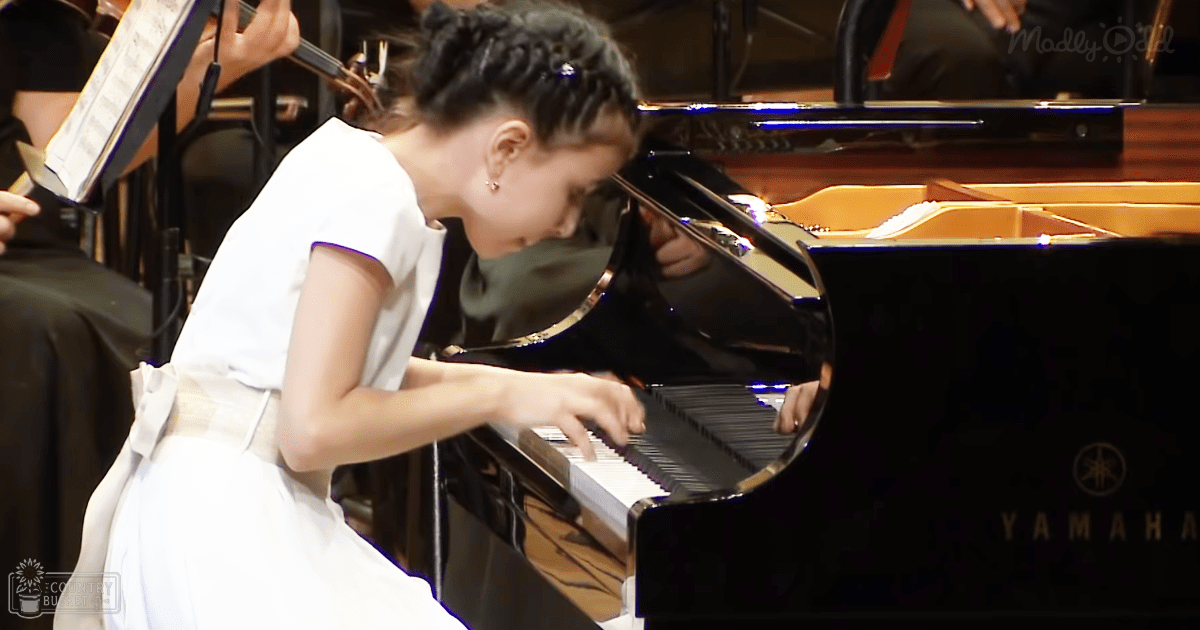 A young child in a white dress plays piano on stage, surrounded by sheet music and other musicians.