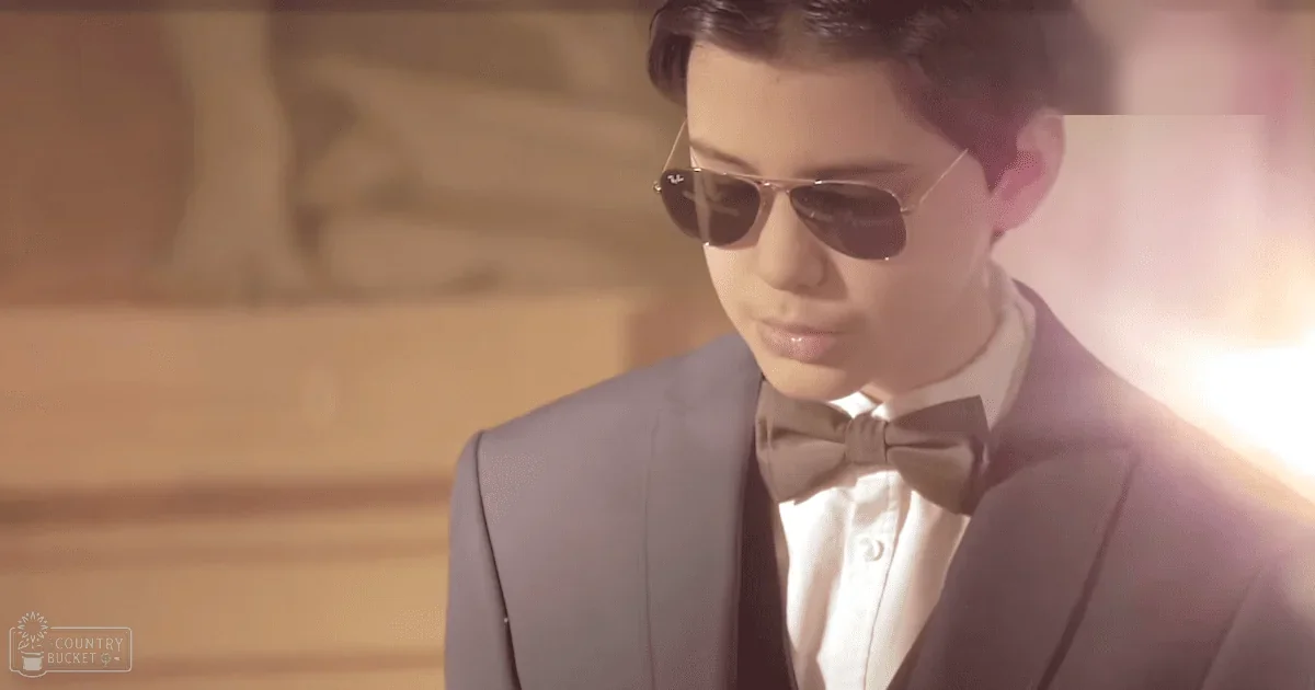 A young adult male wearing a formal light gray suit with a white collared shirt and dark brown bow tie, set against a warm-toned indoor environment with soft lighting.
