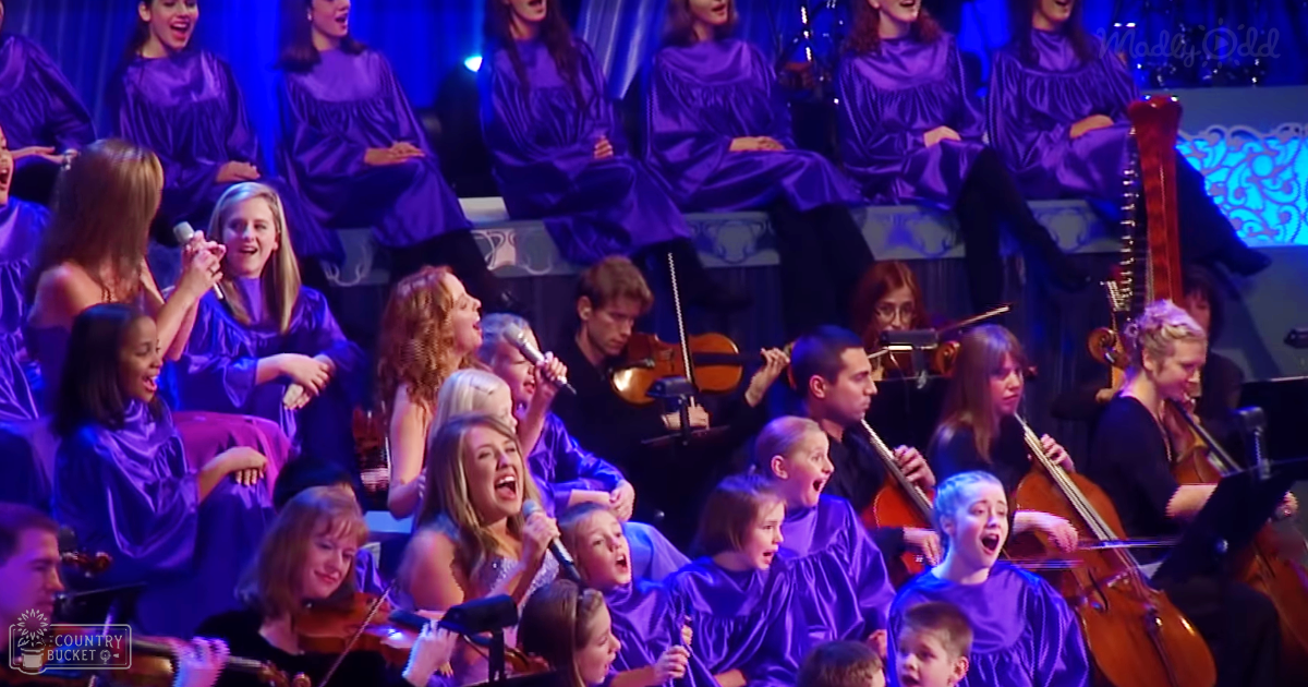 A choir performance with children, adolescents, and adults in matching purple robes held indoors under stage lighting during what appears to be an evening event.