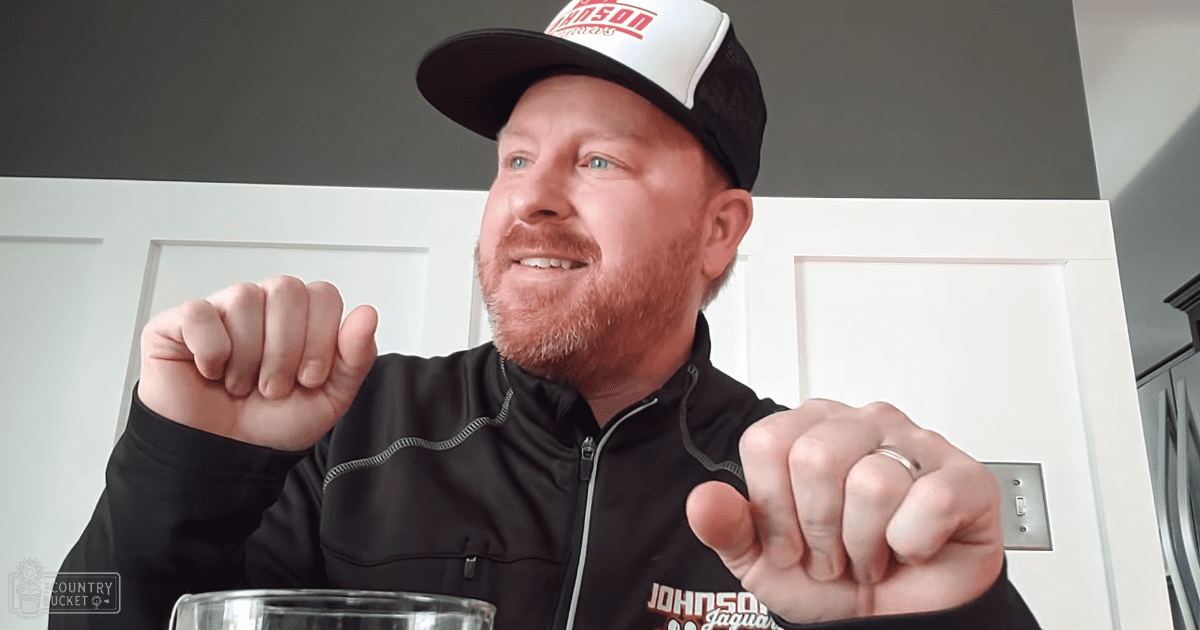 A middle-aged adult male wearing a black zip-up jacket with 'JOHNSON' printed in red letters on his baseball cap stands indoors, with a wedding band visible. The setting includes kitchen cabinetry and natural daylight.