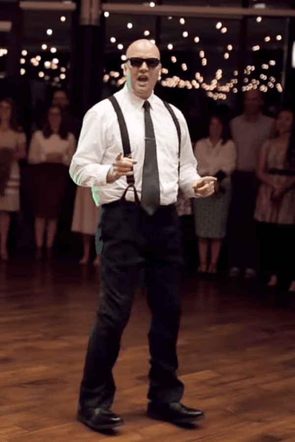 Adult male in a formal white dress shirt with black suspenders stands among middle-aged to older adult females at an indoor event with string lights, suggesting a festive evening gathering.