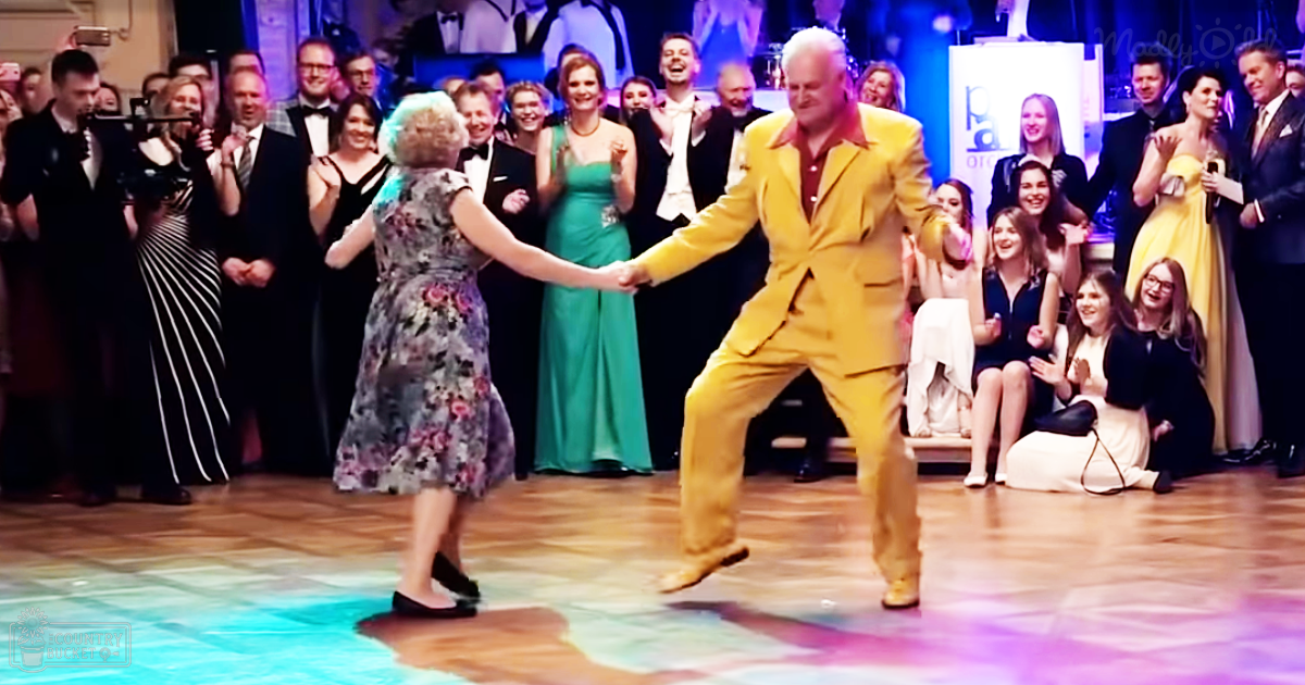 An adult male singer wearing a bright yellow suit dances with an adult female middle-aged dancer in a floral dress at an indoor ballroom event under evening lighting, surrounded by formal attire-clad attendees. A partially visible sign reading 'Molly' is in the background.