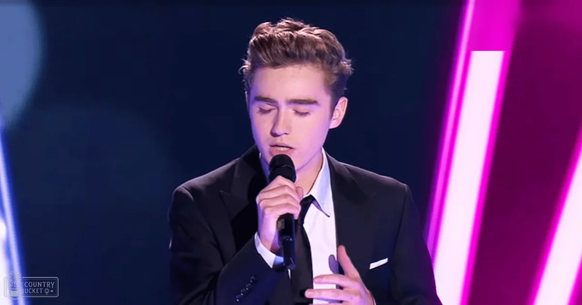 A young adult male in a formal setting, holding a microphone against a backdrop of bright pink and white vertical light strips.