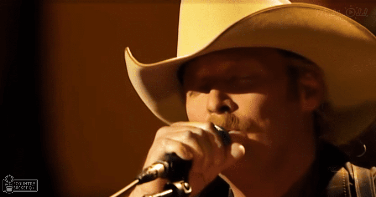 An adult male singer performing indoors, wearing a wide-brimmed cowboy hat with light-colored band and dark shirt with decorative stitching; dimly lit stage setting suggests evening or night time performance space.
