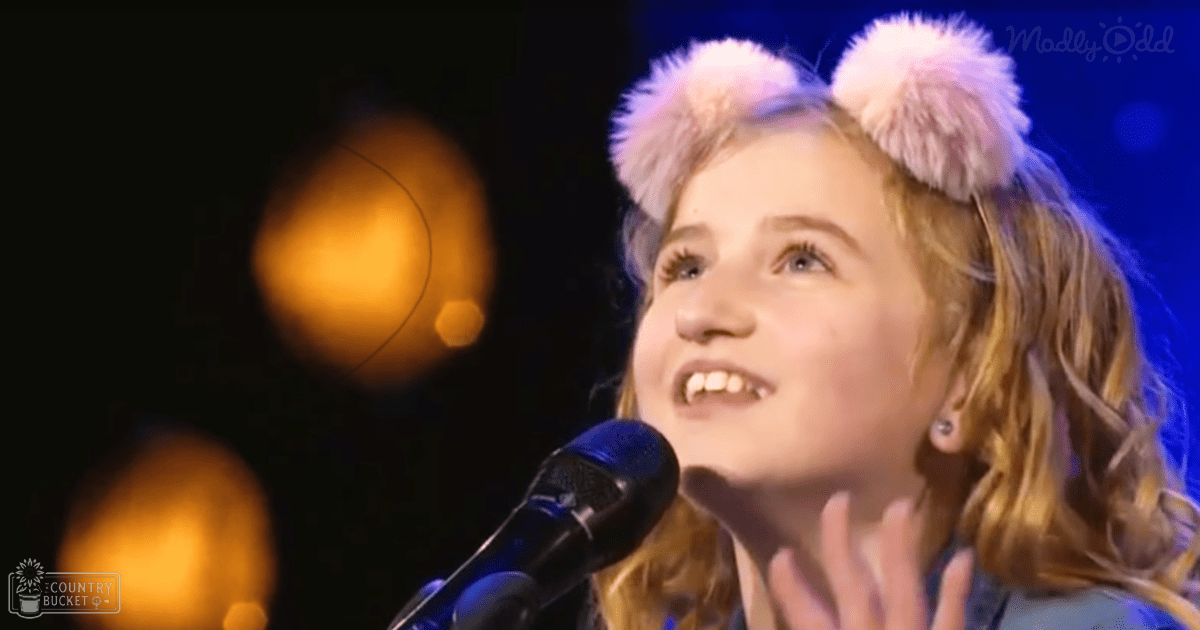 A young female child wearing a blue collared jacket with long sleeves, white fur-like headbands, and small earrings stands on stage at an indoor event, likely performing or speaking into a microphone under stage lighting.