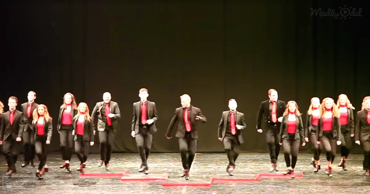 A performance group on stage with a dark backdrop at an indoor theater; adults in black suits and blazers with red ties, white shirts, and black shoes;
