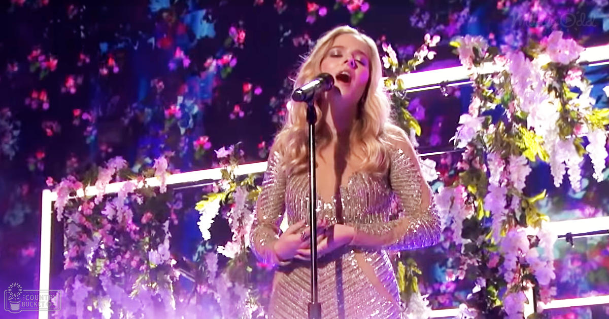 An adult female singer performing indoors, wearing a shimmering sequined dress with floral decorations in the background.
