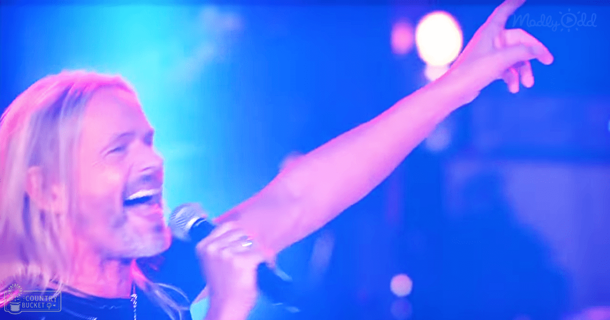 A middle-aged male singer performing at a concert, holding a microphone under stage lighting with blue and white hues, in an indoor music venue.