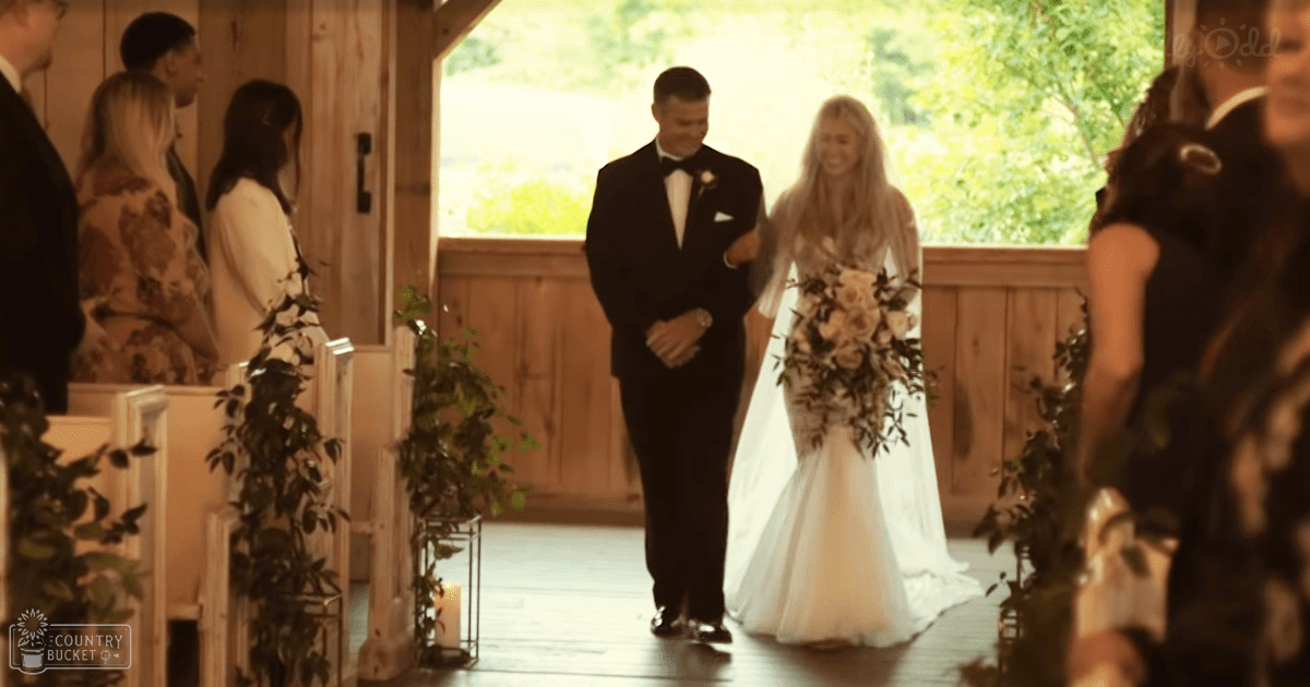 A bride in a white wedding gown walks down an aisle with her father at a rustic barn-style wedding venue during late afternoon, with wooden pews adorned with greenery and small candle holders.