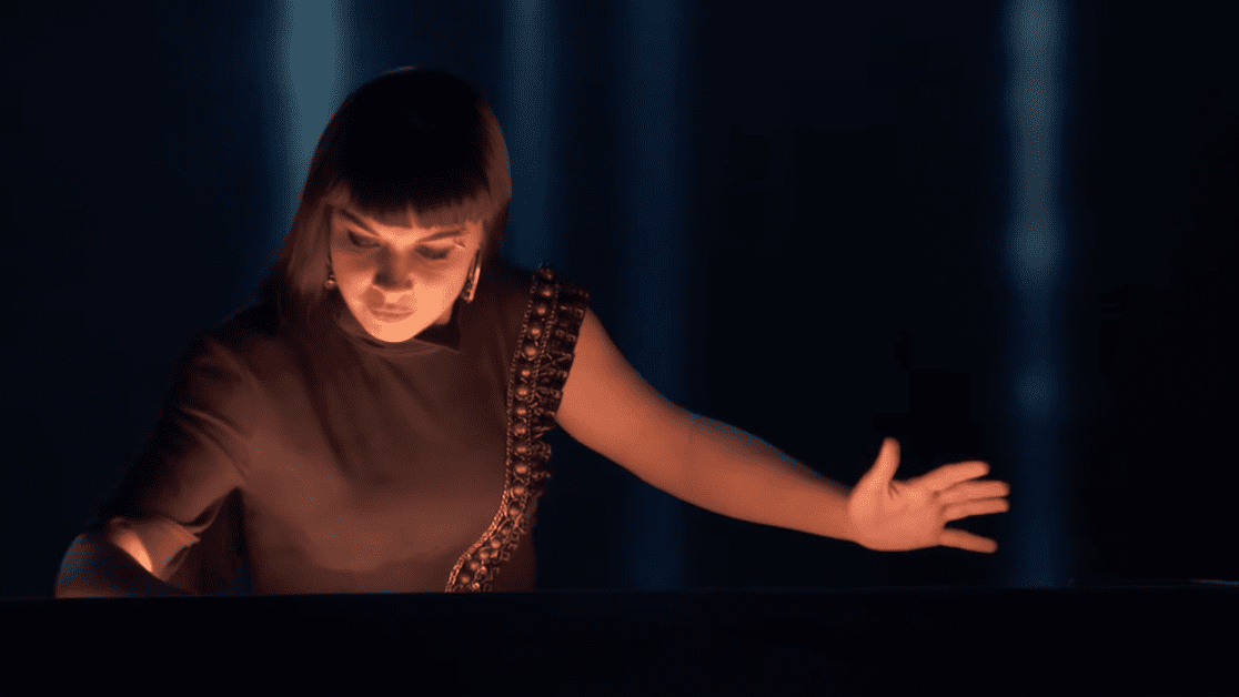 A young adult female in an elegant, sleeveless top with intricate detailing sits at a piano indoors. Dark curtains and dramatic lighting create an intimate atmosphere.