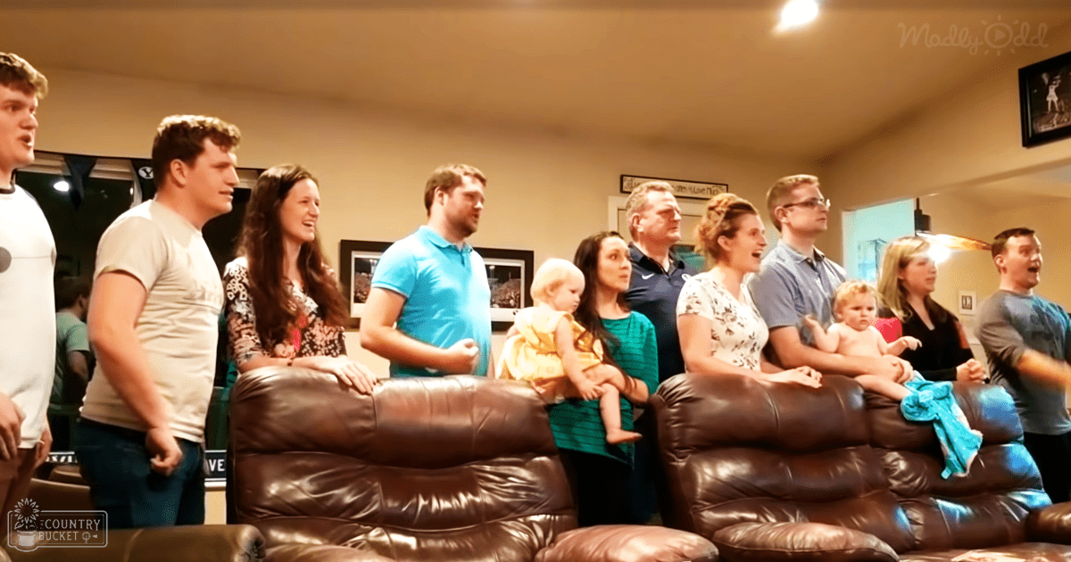 A family gathering in a cozy living room with adults and children indoors. Brown leather sofas, framed wall decor, and indoor lighting create a homey atmosphere.