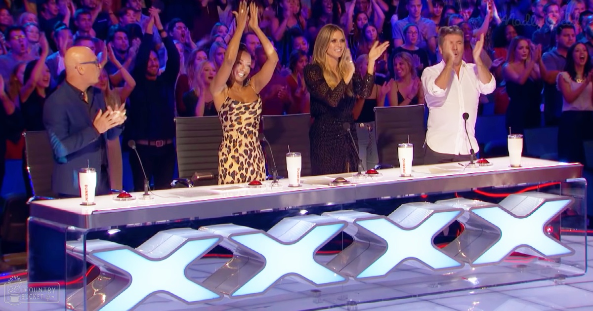 A panel judging 'The X Factor' talent show indoors, with judges and audience clapping and cheering under stage lighting.