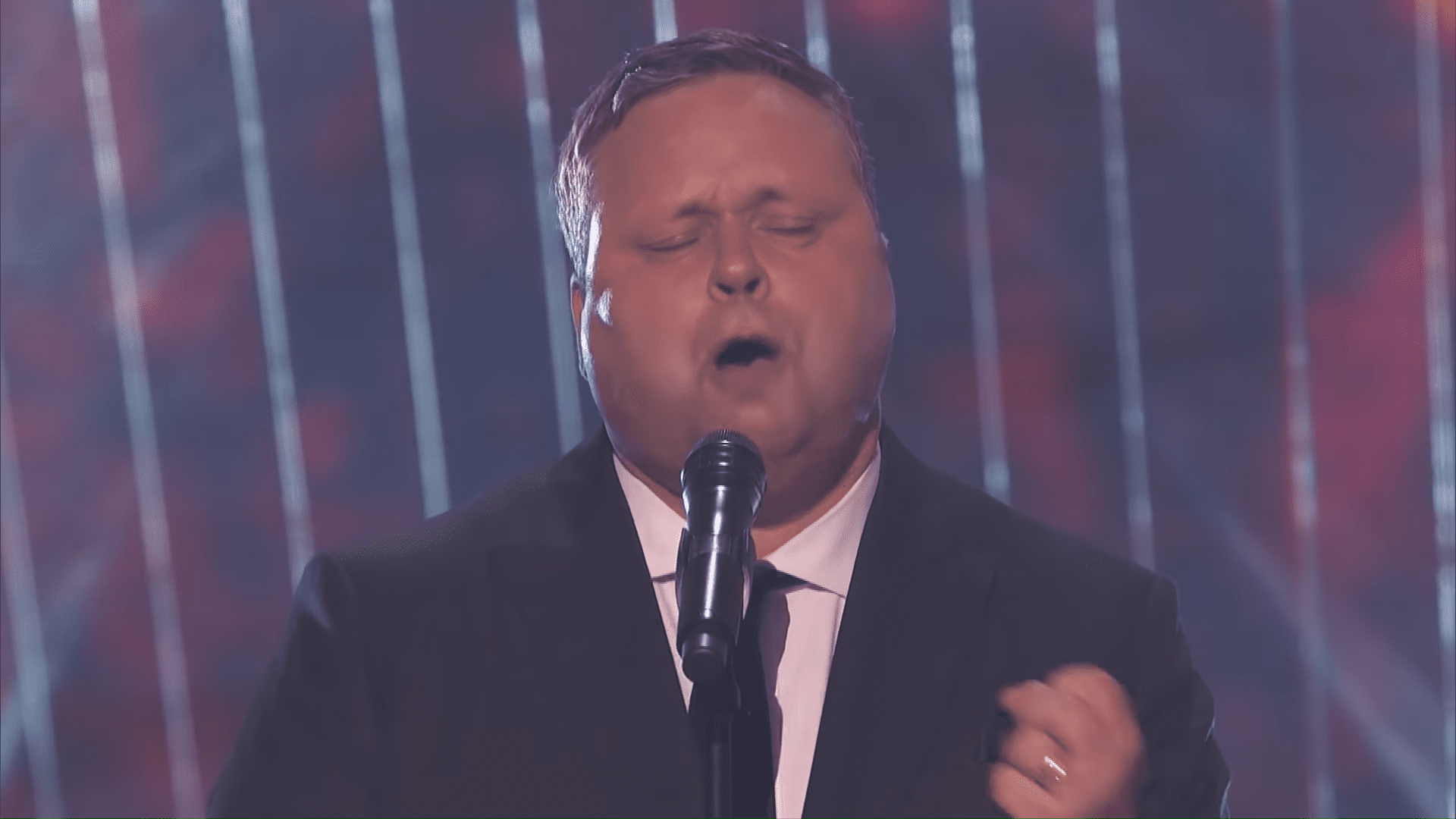A middle-aged man singing on a dimly lit indoor stage with professional lighting and dark vertical light strips behind him.