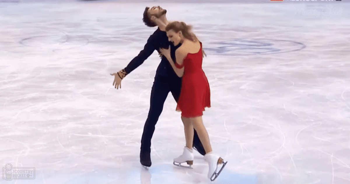 A male figure skater in a black outfit with dark pants stands on an indoor ice rink alongside his female partner wearing a red sleeveless dress, both wearing white figure skates under artificial evening lighting.