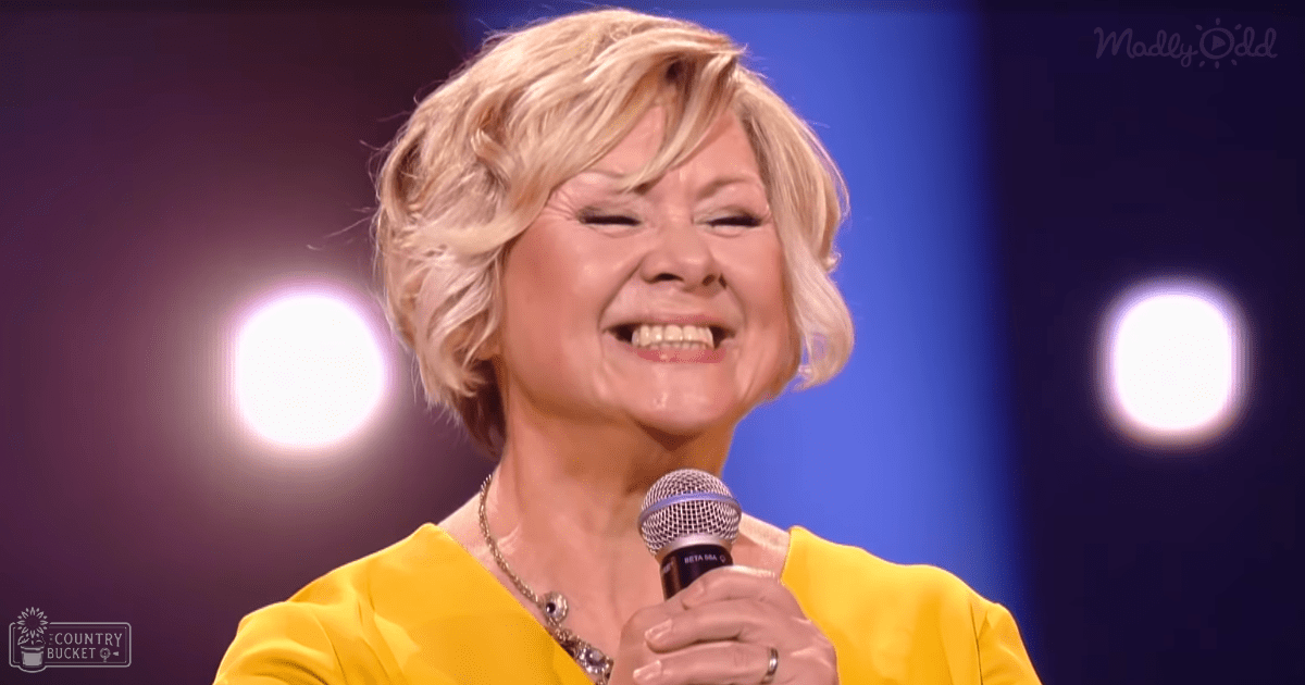 A middle-aged adult female wearing a bright yellow relaxed-fit long-sleeve top with a round neckline and a necklace with a pendant performs on stage under artificial lighting, holding a 'BETA 58A' microphone.