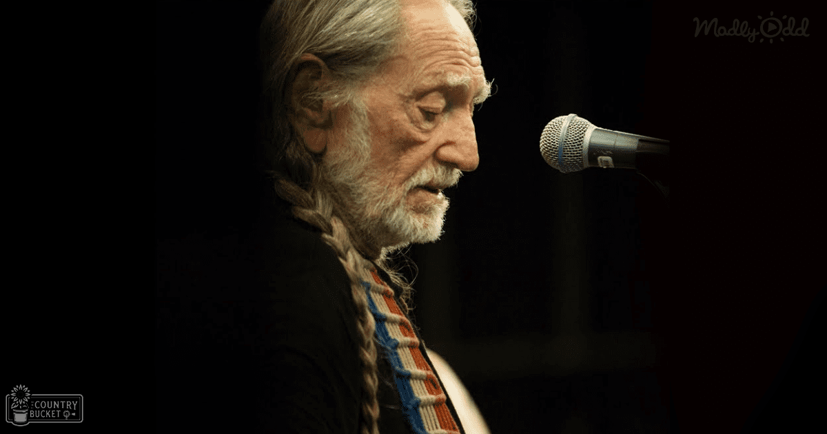 A middle-aged man wearing a black, striped long-sleeved shirt or jacket with red and blue patterns performs on stage in an indoor venue under dim lighting.