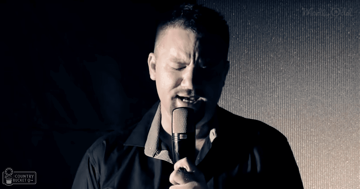 A young adult male singer in a relaxed modern casual style, wearing a dark-colored jacket over a plaid shirt, holding a microphone during a performance with controlled stage lighting.