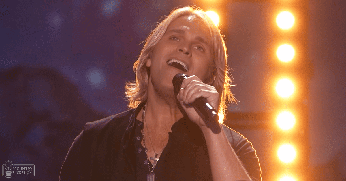 An adult male singer performing on a dimly lit indoor concert stage, wearing a dark button-up shirt with rolled sleeves and a black vest over it, holding a microphone with bright yellow-orange stage lights in the background.