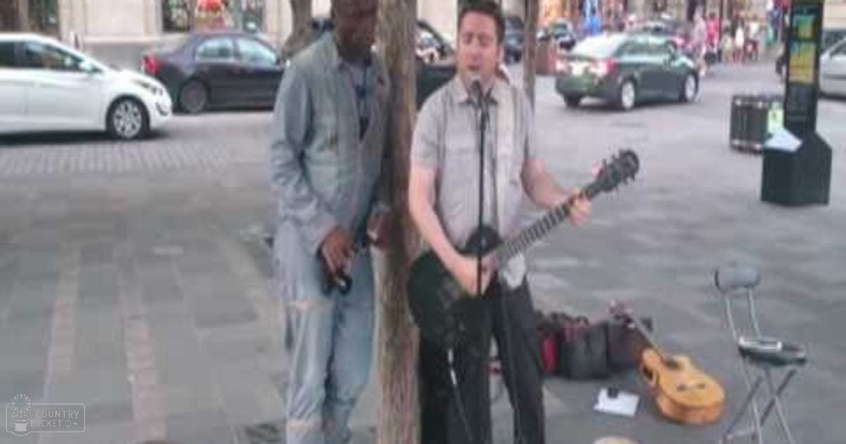 Urban street performer playing guitar with a young adult companion, spring or summer day in an urban setting with parked cars and pedestrians.