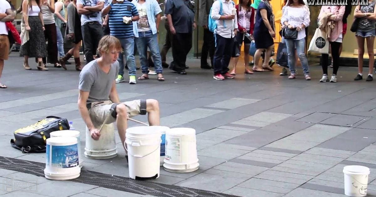 A young adult male street performer plays drums with buckets in an urban plaza surrounded by a casual audience on a sunny day.