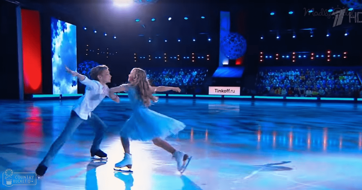 A child ice skater wearing a white long-sleeved shirt and dark pants performs alongside another child in a light blue tutu dress on an indoor rink during what seems to be a skating competition or performance.