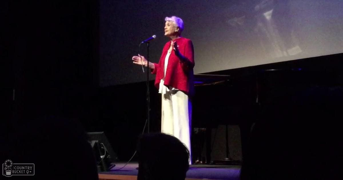 An older adult female performer wearing a red textured blazer and white tailored pants stands on stage with a microphone stand to her left, in an indoor theater or concert hall setting.