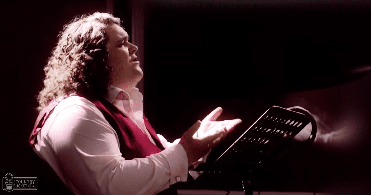 Young adult male in a recording studio wearing a white dress shirt with long sleeves and a red vest; sheet music on a music stand.
