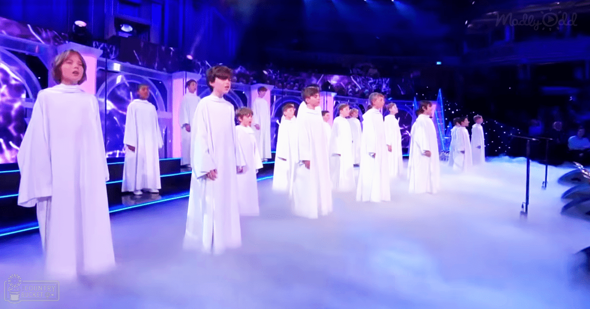 A choir of children in white robes performing on stage with blue and purple lighting, likely in a concert hall or theater setting.