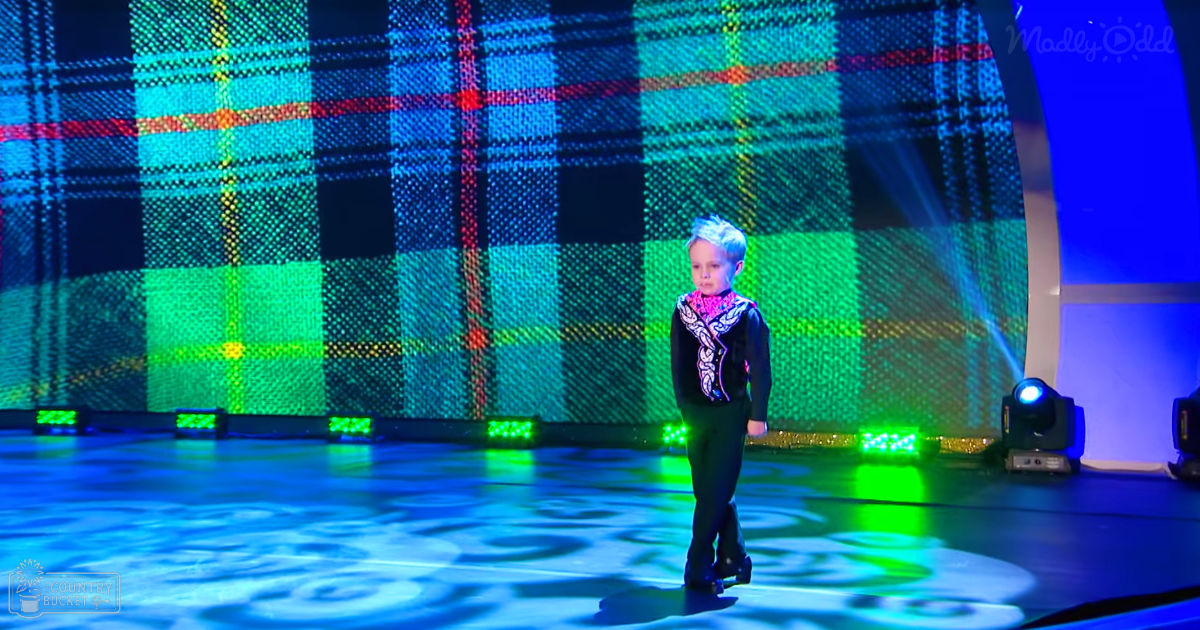 A child wearing a black jacket with a white patterned scarf stands on stage under colorful lights during an indoor event or performance.