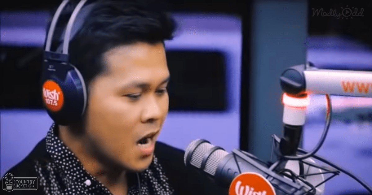 A young adult male in a recording studio, wearing a black jacket with white polka dots and black headphones marked 'Wish 107.5', with a red-lit microphone base and a monitor screen in the background.