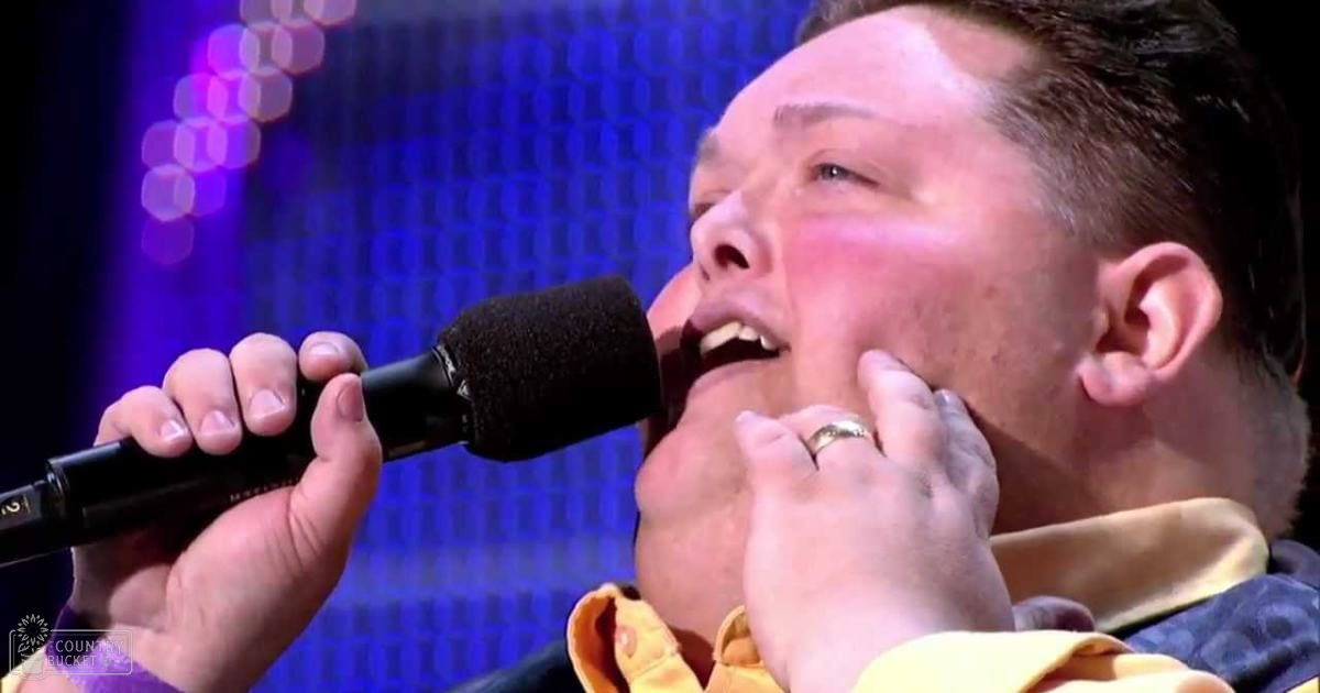 A middle-aged adult male in a regular-fit, collared button-up shirt with long sleeves, wearing a wedding band, sings into a microphone at an indoor event with stage lighting visible behind him.