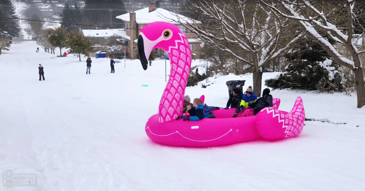 Children play on a large pink inflatable flamingo sled in a snowy residential street, with snow-covered trees and houses in the background.