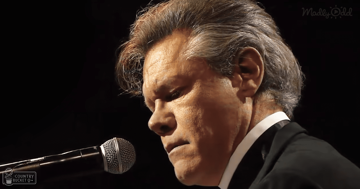 Middle-aged adult male in a formal black suit jacket, white shirt with no visible tie, speaking or singing into a microphone against a dark indoor backdrop.