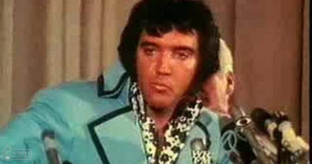 A middle-aged man with dark, upward-styled hair in a light blue suit jacket with bold black and white patterned shirt indoors.